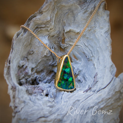 Free-form cut triangular shaped opal with tapered besel setting in 14K yellow gold. No bail needed with the chain passing through a whole included in the besel setting. Necklace is hanging off a light-grey grained piece of driftwood.