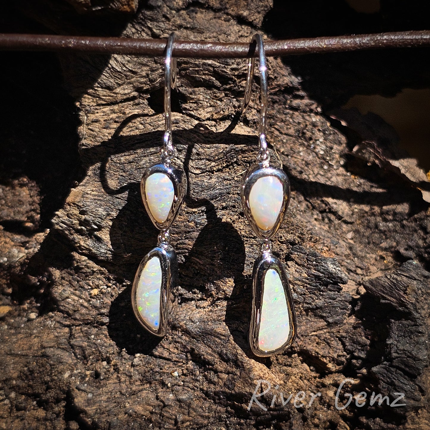 Two smaller light opals at the top are tear drop shaped and the bottom ones are free-form cut. Eyelet connections between the two besel set gems and hook connection. White opals contrast against the dark wood grain background.