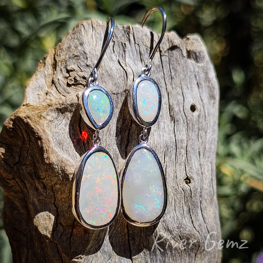 Two multi-coloured opals in each French hook earring. Opals are connected with eyelets between each other and the hook. Hooks are presented against the light coloured weathered drift wood.