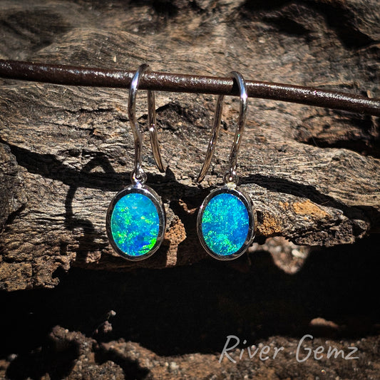 Vivid blue-green opal in the cabochon cut opal French hook earrings. Eyelet hooks connect the besel set opal to the hook of the earring. Opals stand out against the dark wood grain background.