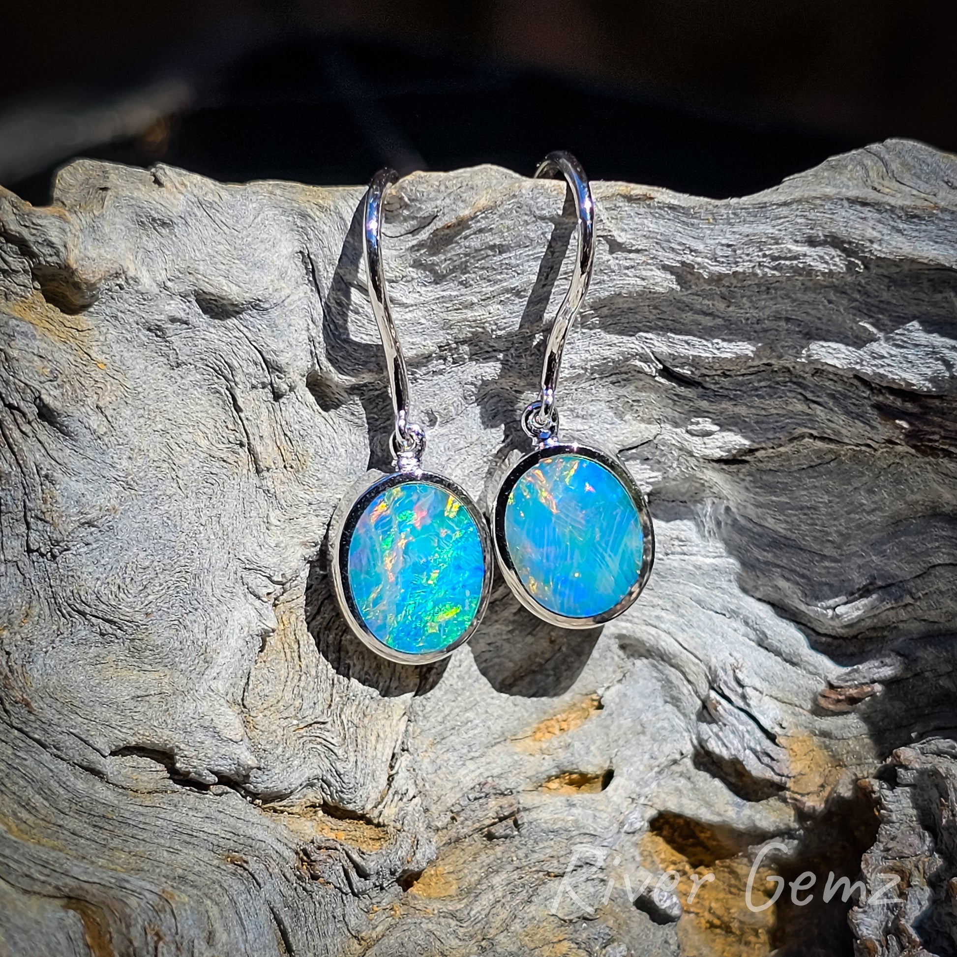 Multi-coloured opals with flashes of the rare red colour in both cabochon cut opals. Eyelet connection between besel set opal and the earring hook. Light grain wood background contrasts with the dark hues of these Crystal opals.