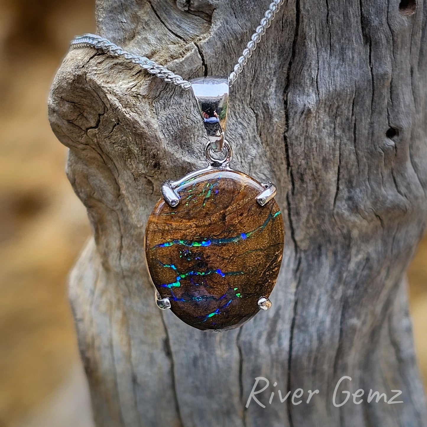 Several horizontal veins of blue-grey colour in the approximately oval shaped boulder matrix opal. The brown of the ironstone host rock contrasts with the light coloured drift wood from which the necklace hangs. 