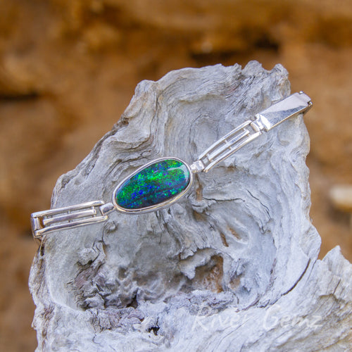 Approximately oval shaped opal besel set in the silver bracelet. The bracelet is drooped over a light-grey piece of drift wood.