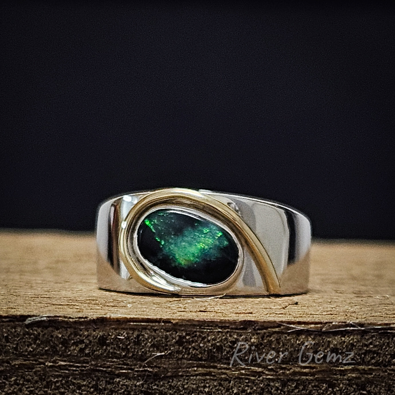 Second front view of the oval shaped opal which is slightly angled in the ring shown  sitting on wooden plank and deep black background to the image.