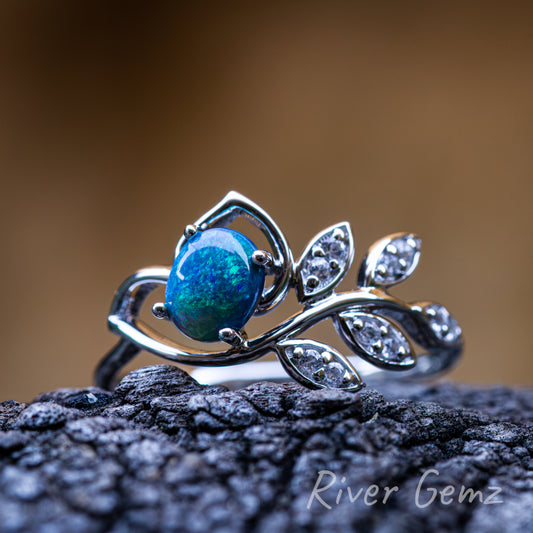 Oval shaped opal held securely by 4 claws in silver ring which has 5 white topaz adorned branch leaves on the shank of the ring. Ring shown sitting on dark grained drift wood and a blurred brown toned back ground.