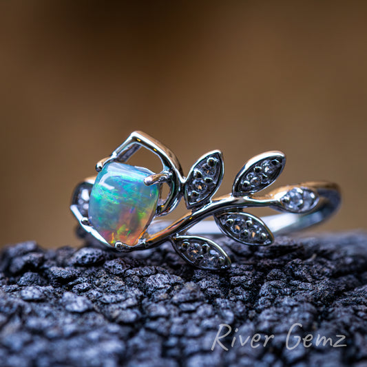 Crystal opal cut to form a four-sided shape held securely by 4 claws in the silver ring. Five leaves adorned with white topaz on the shank of the ring. Ring shown resting on a dark textured piece of drift wood and a blurred light brown backdrop.