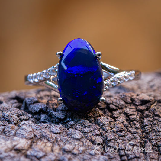 Front view of dark blue oval shaped black opal held securely by 4 claws in the silver ring with topaz stones each side of the opal. Ring is positioned on a dark grained piece of drift wood and burnt orange back drop.