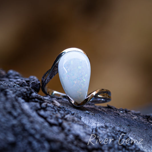 White opal partial besel set in a sophisticated modern styled silver ring. Ring sits on a dark grained piece of weathered drift wood and the photo back drop is a blurred burnt orange.