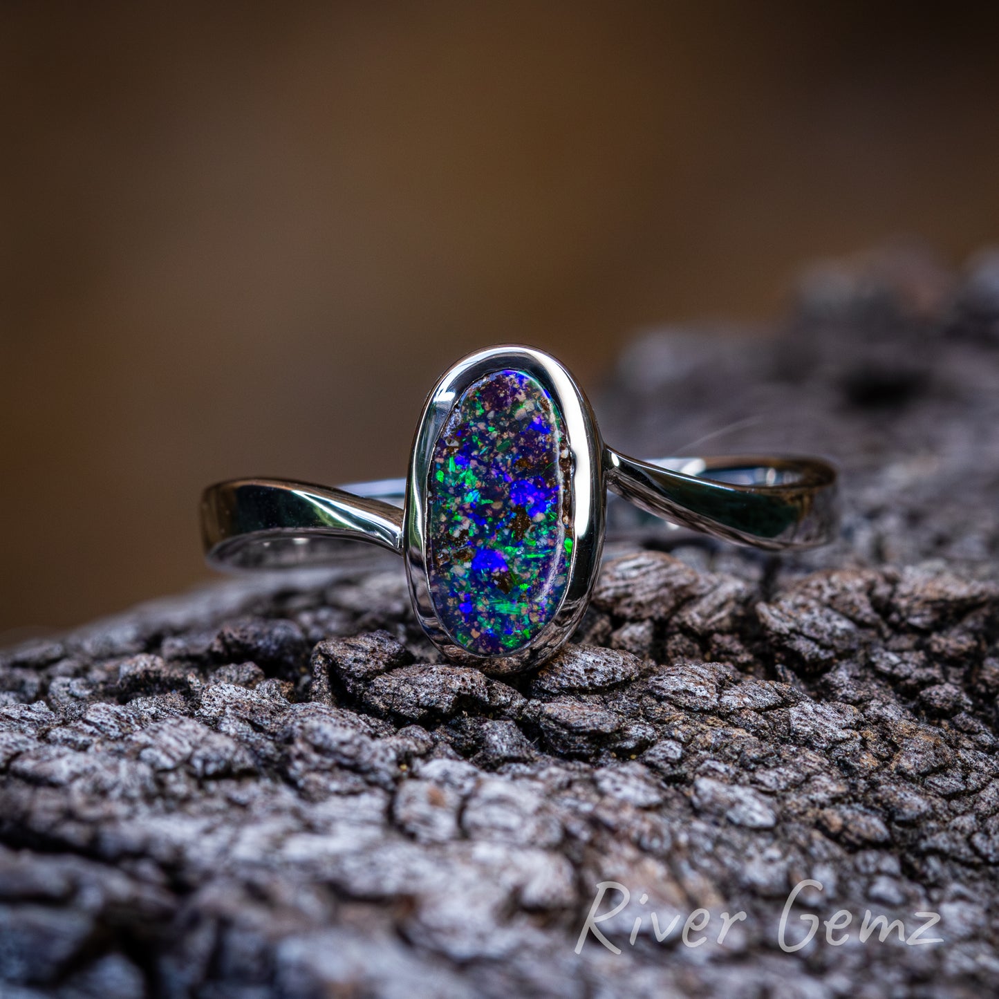 Free form cut approximately oval shaped colourful opal in a simple but elegant silver setting. The ring sits on a dark grained piece of drift wood and the silver contrasts against the smokey orange blurred back drop.