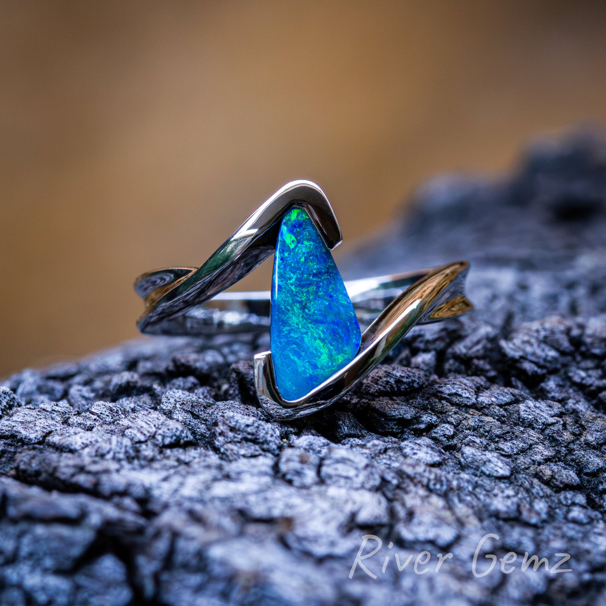 Free-form cut triangular shaped blue-green boulder opal in a modern sterling silver ring design. Ring is shown on dark grained drift wood with burnt orange backdrop