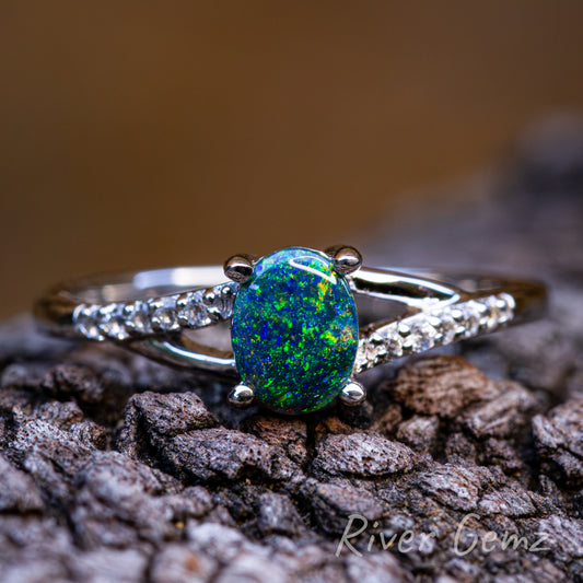 Cabochon mostly blue-green opal held securely with 4 claws in silver ring. Ring sits on a dark grained piece of driftwood and burnt orange backdrop. 