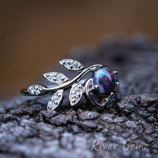 Multi-coloured cabochon black opal held securely with 4 claws in sterling silver ring. Setting includes 5 leaves with topaz stones. Ring rests on a dark grained drift wood and blurred backdrop.