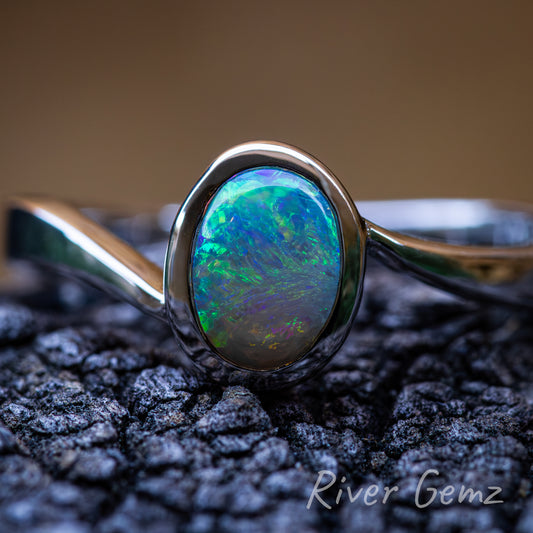 Swirls of colour in the cabochon cut crystal opal besel set in silver ring. Ring sits on dark grained piece of driftwood with a light brown back ground.