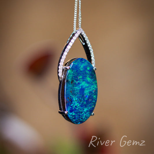 Oval shaped dark blue and green solid opal held securely by 4 claws in the white gold setting. A "crown" of diamonds completes the necklace design. Blurred burnt orange back drop used in the photo. This slightly side on view shows the ironstone of the boulder opal and also the thickness of the opal.