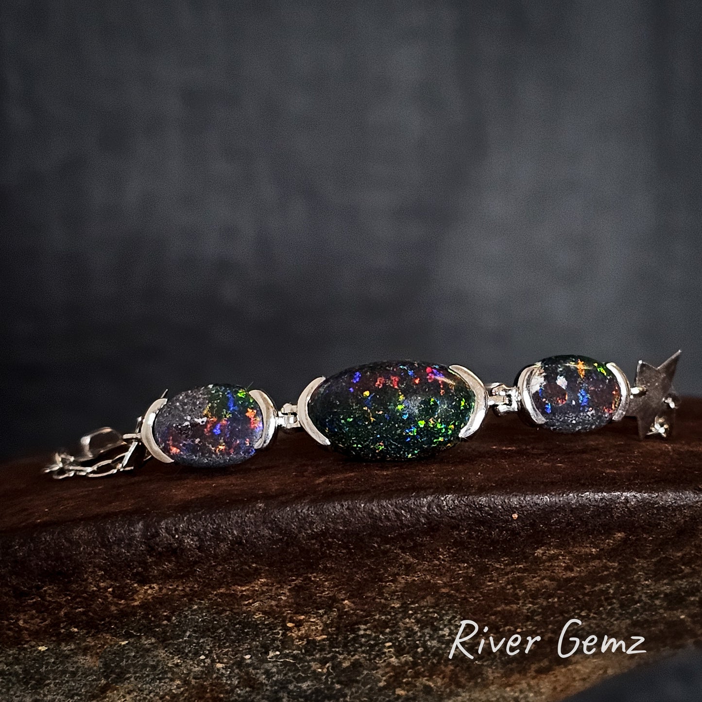 Silver bracelet with three colourful oval shaped matrix opals on a dark background, featuring the brand 'River Gemz'.