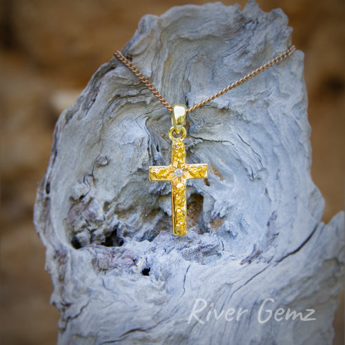 Gold nugget pendant is shown on a light-grey weathered piece of driftwood. The pendant includes a gold bail which passes through an eyelet at the top of the cross.