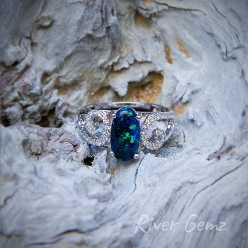 Calibrated oval shaped dark opal with blue & green colours together with 2 swirls of small diamonds each side in this modern designed white gold ring. 