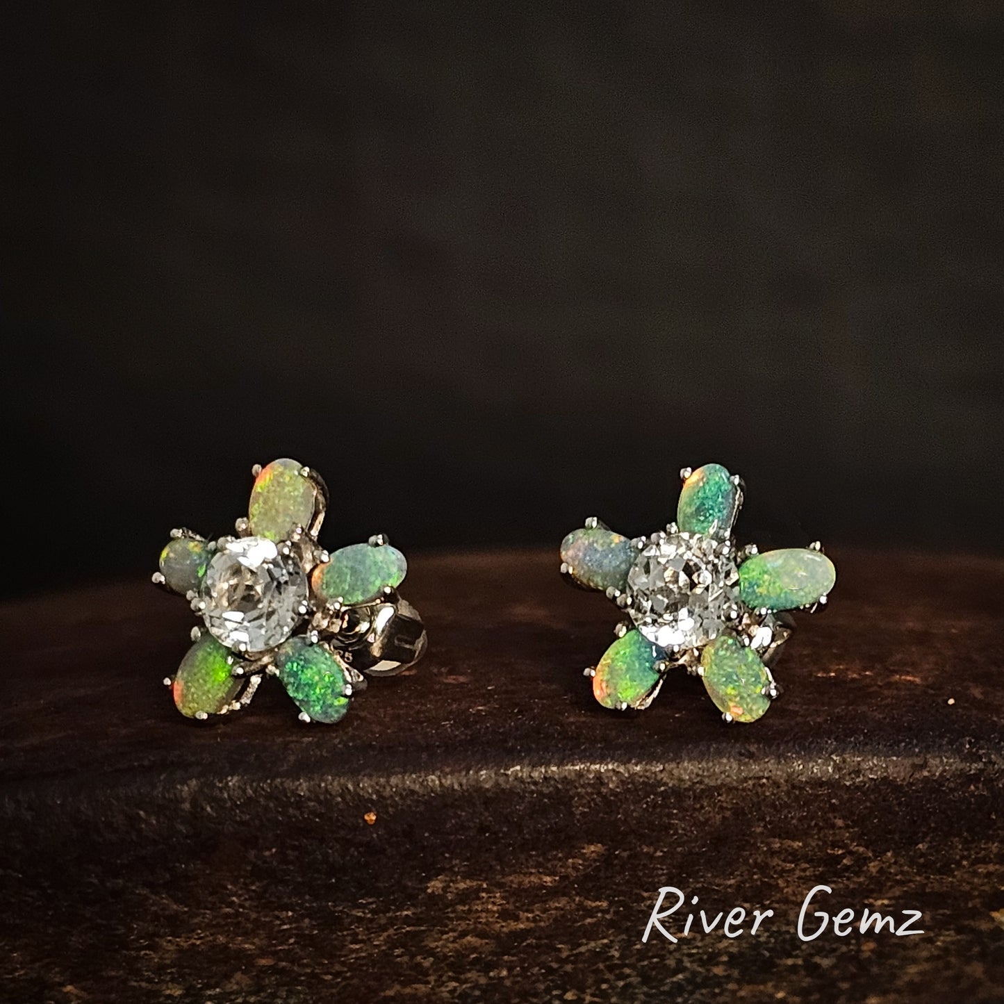 Pair of flower-shaped earrings with green gemstones on a dark surface, branded 'River Gemz'.