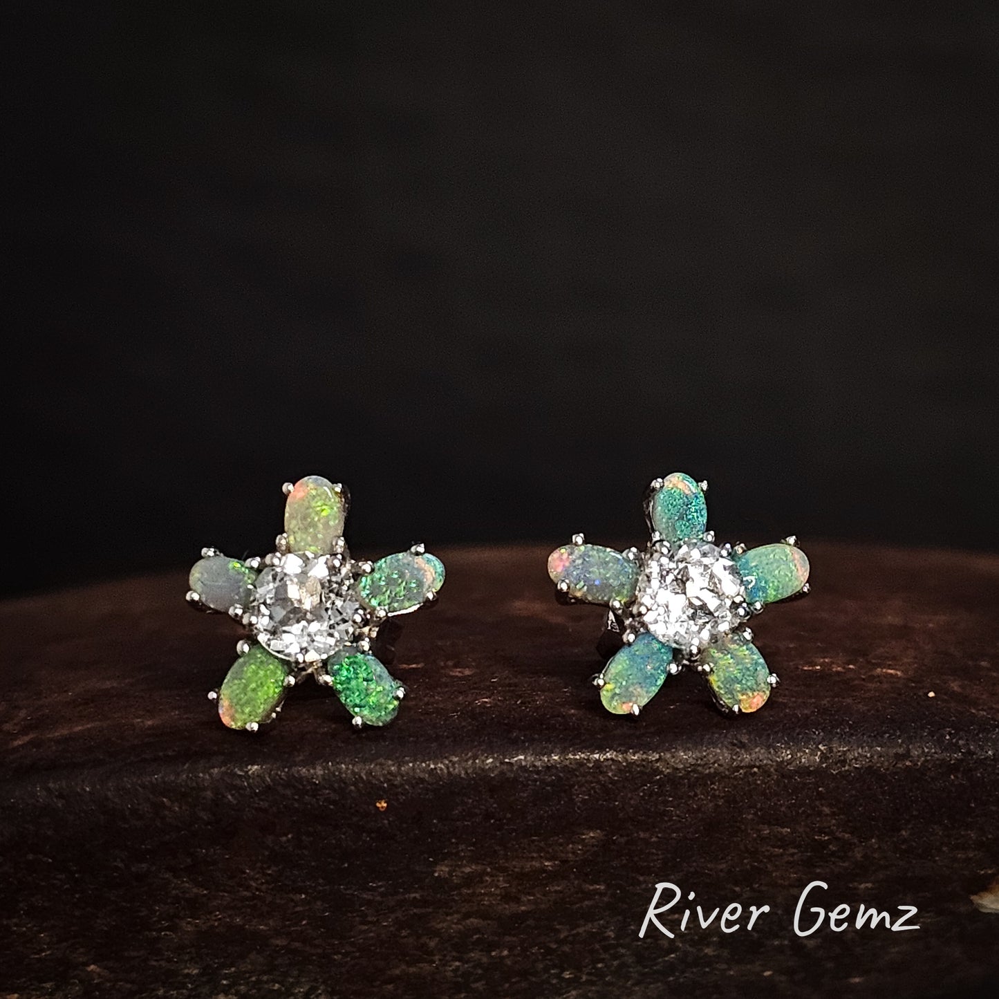 Pair of flower-shaped earrings with green and blue opal stones on a dark background, branded 'River Gemz'.