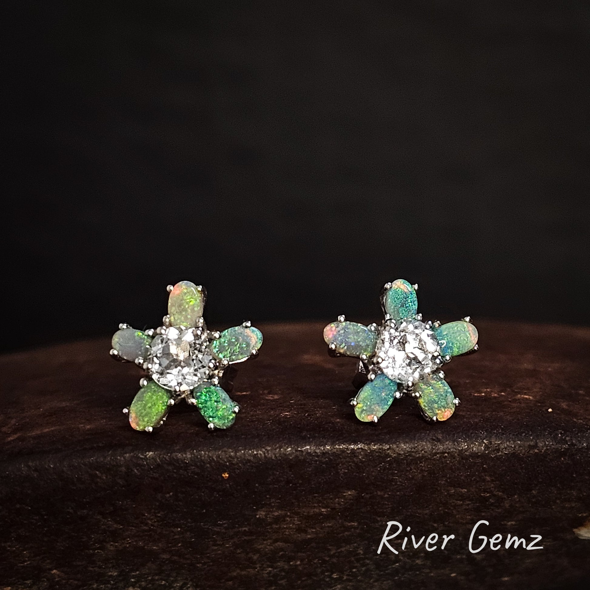 Pair of flower-shaped earrings with green and blue opal stones on a dark background, branded 'River Gemz'.