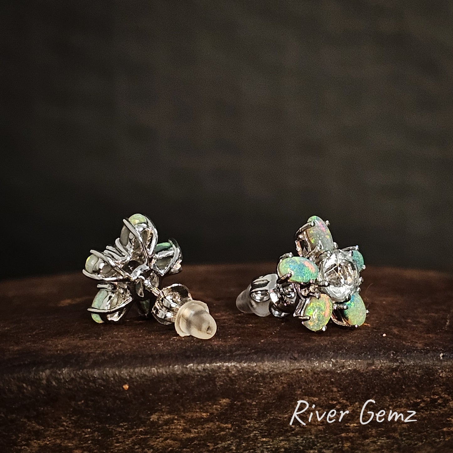 Pair of earrings with 5 claw set light opals and a central white topaz stone on a cast iron surface, branded 'River Gemz'.