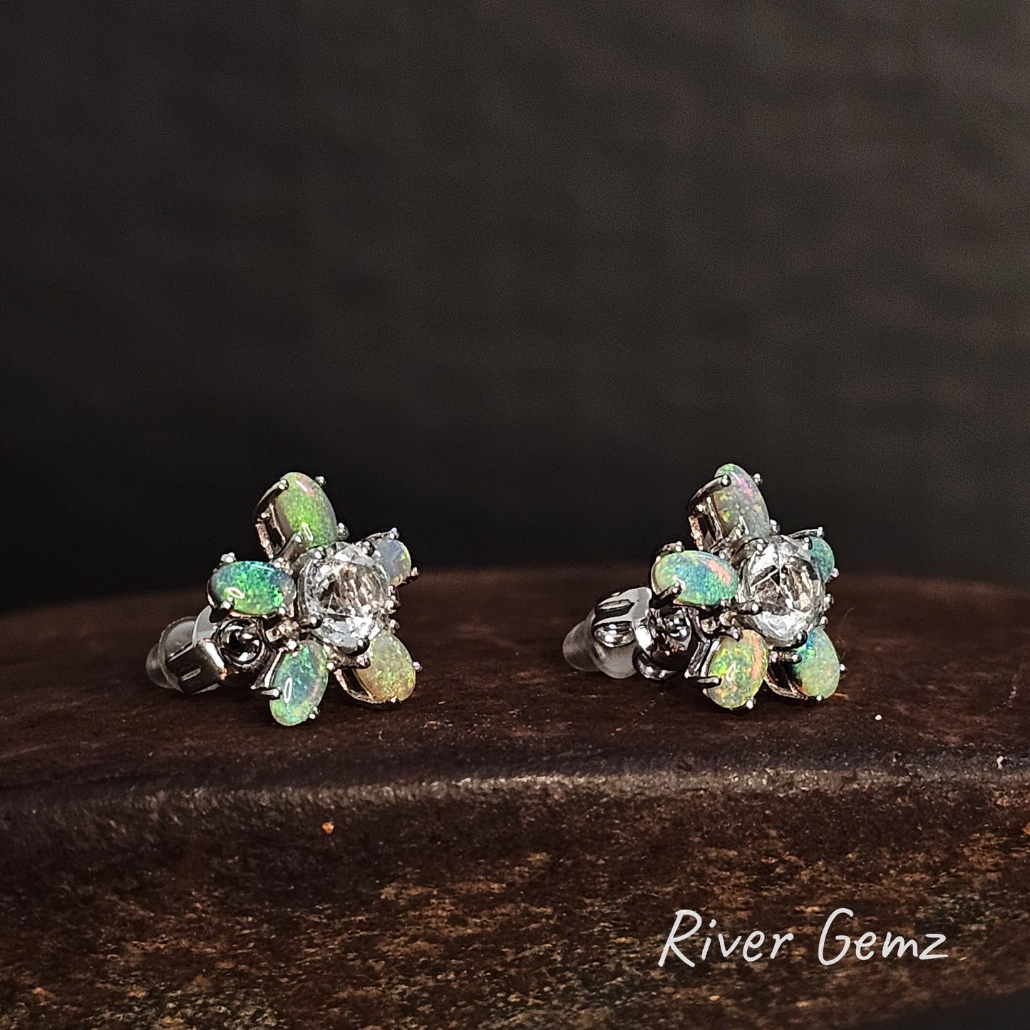 Pair of silver earrings with 5 oval shaped green crystal opals around a white topaz stone on a dark surface, branded 'River Gemz'.
