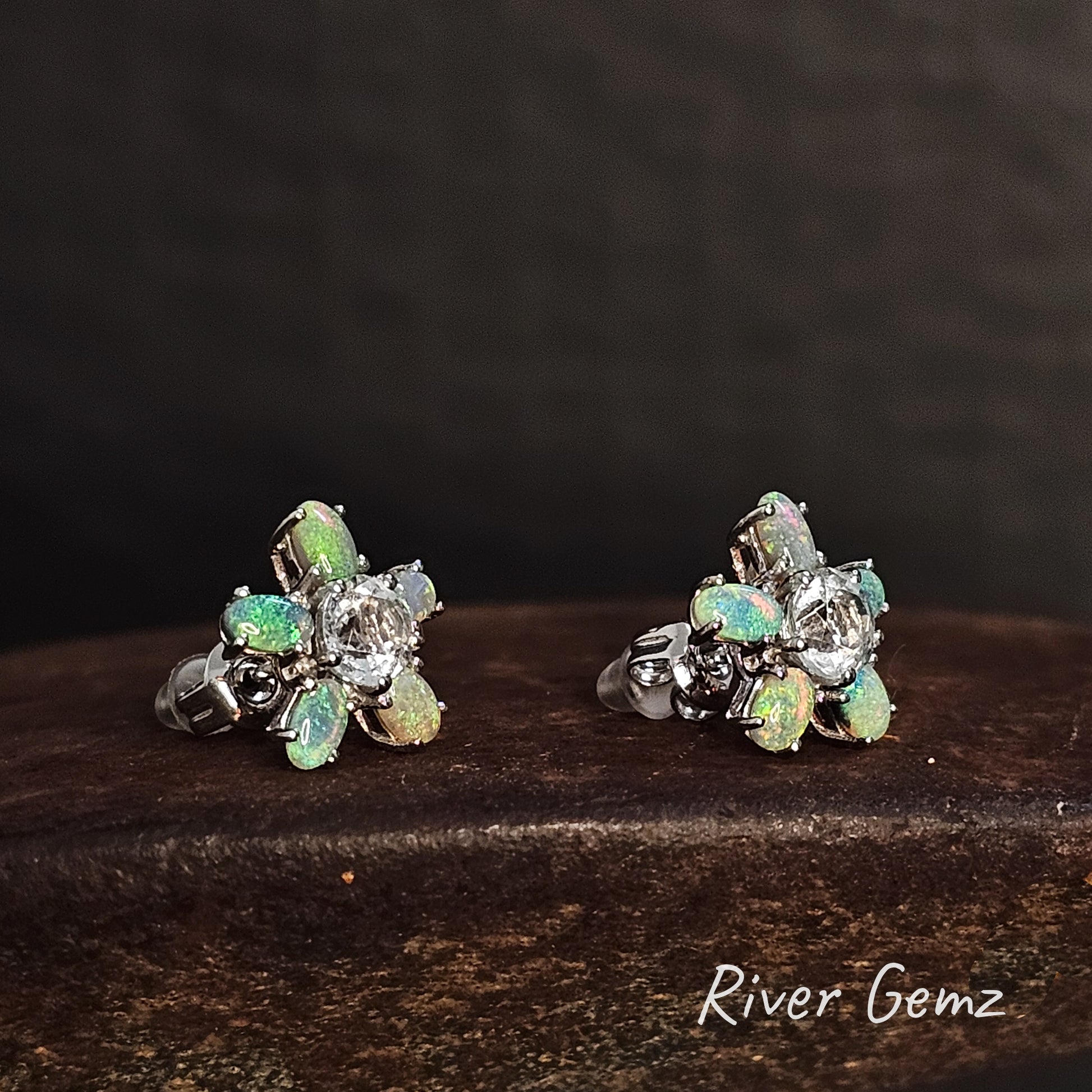 Pair of silver earrings with 5 oval shaped green crystal opals around a white topaz stone on a dark surface, branded 'River Gemz'.