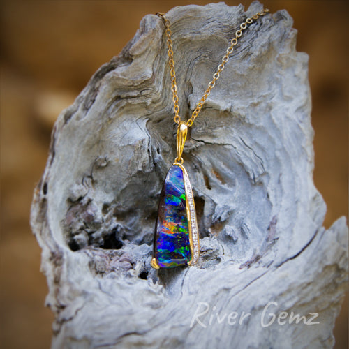 Approximately triangular shaped vividly coloured boulder opal claw and besel set in 18K yellow gold. Tapered bail through the eyelet completes the pendant. Dark colours of opal contrast with the light-grey driftwood it is photographed against.