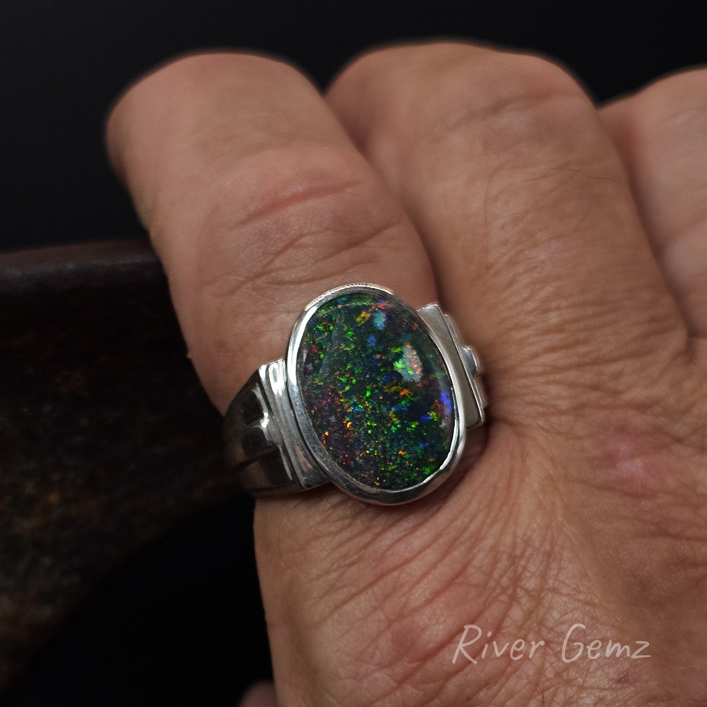 Ring with a colorful opal on a person's finger, branded 'River Gemz'.