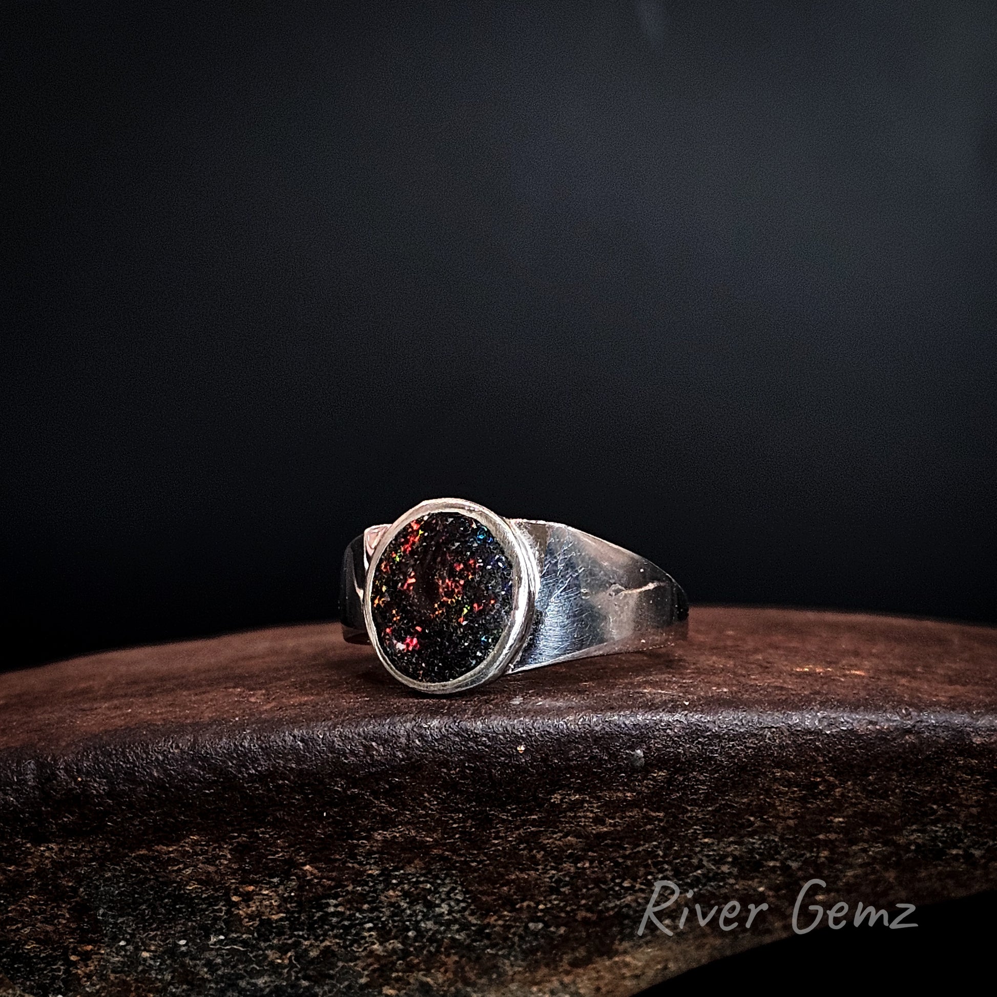 Multi-coloured matrix opal full bezel set in sterling silver ring on rusty coloured surface with 'River Gemz' branding.