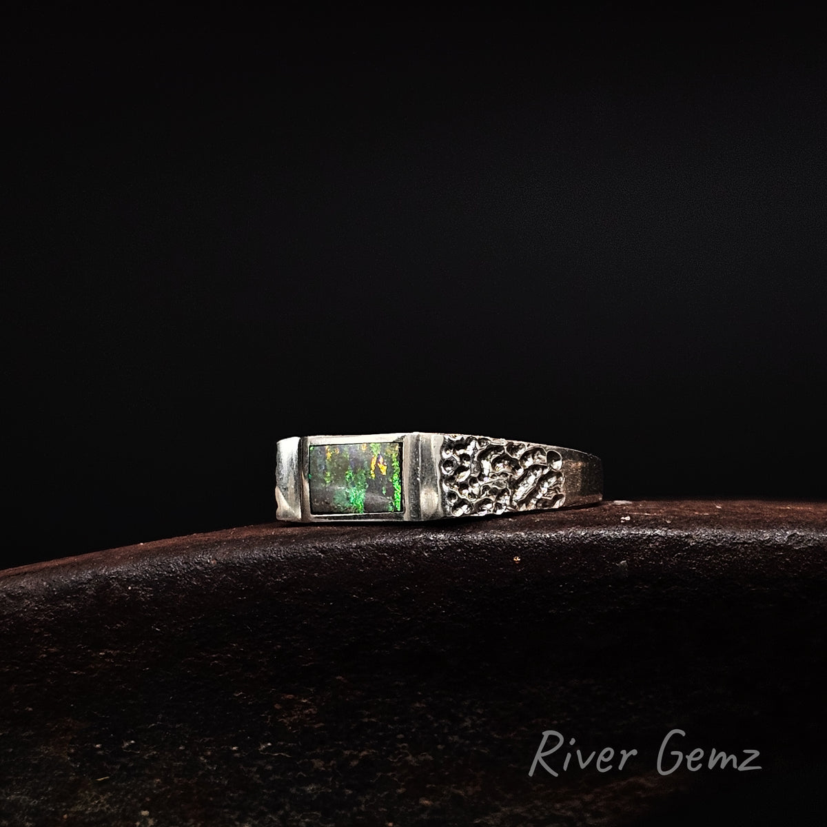 A sterling silver ring featuring a rectangular green opal gemstone on one side and a textured, hammered pattern on the other, resting on a dark brown surface against a black background. The 'River Gemz' brand name is visible in the bottom right corner.