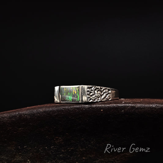 A sterling silver ring featuring a rectangular green opal gemstone on one side and a textured, hammered pattern on the other, resting on a dark brown surface against a black background. The 'River Gemz' brand name is visible in the bottom right corner.