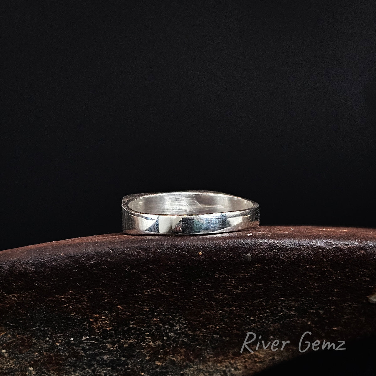 Rear view of silver ring showing the silver backing behind the inset opal, resting on a dark brown textured surface against a solid black background. The brand name 'River Gemz' is visible as a watermark in the bottom right corner.