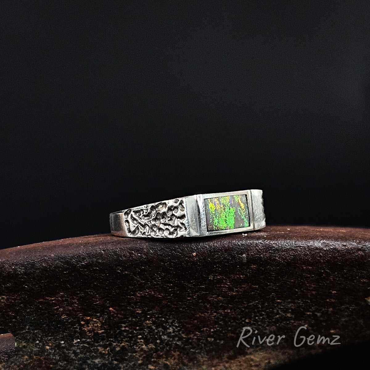 A silver ring with an engraved band and a square green opal inset, resting on a dark wooden surface against a black background. The watermark 'River Gemz' is visible in the bottom right corner.