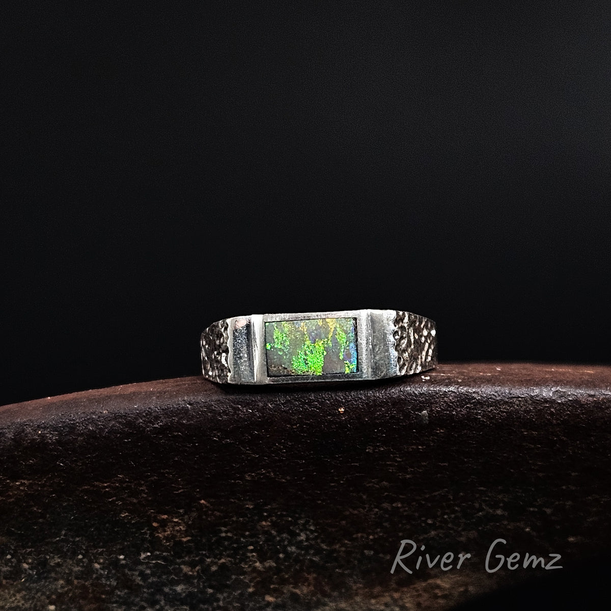 A silver-toned ring featuring a rectangular green iridescent opal inlay, displayed on a dark textured surface against a black background. The brand name 'River Gemz' is visible in the bottom right corner.