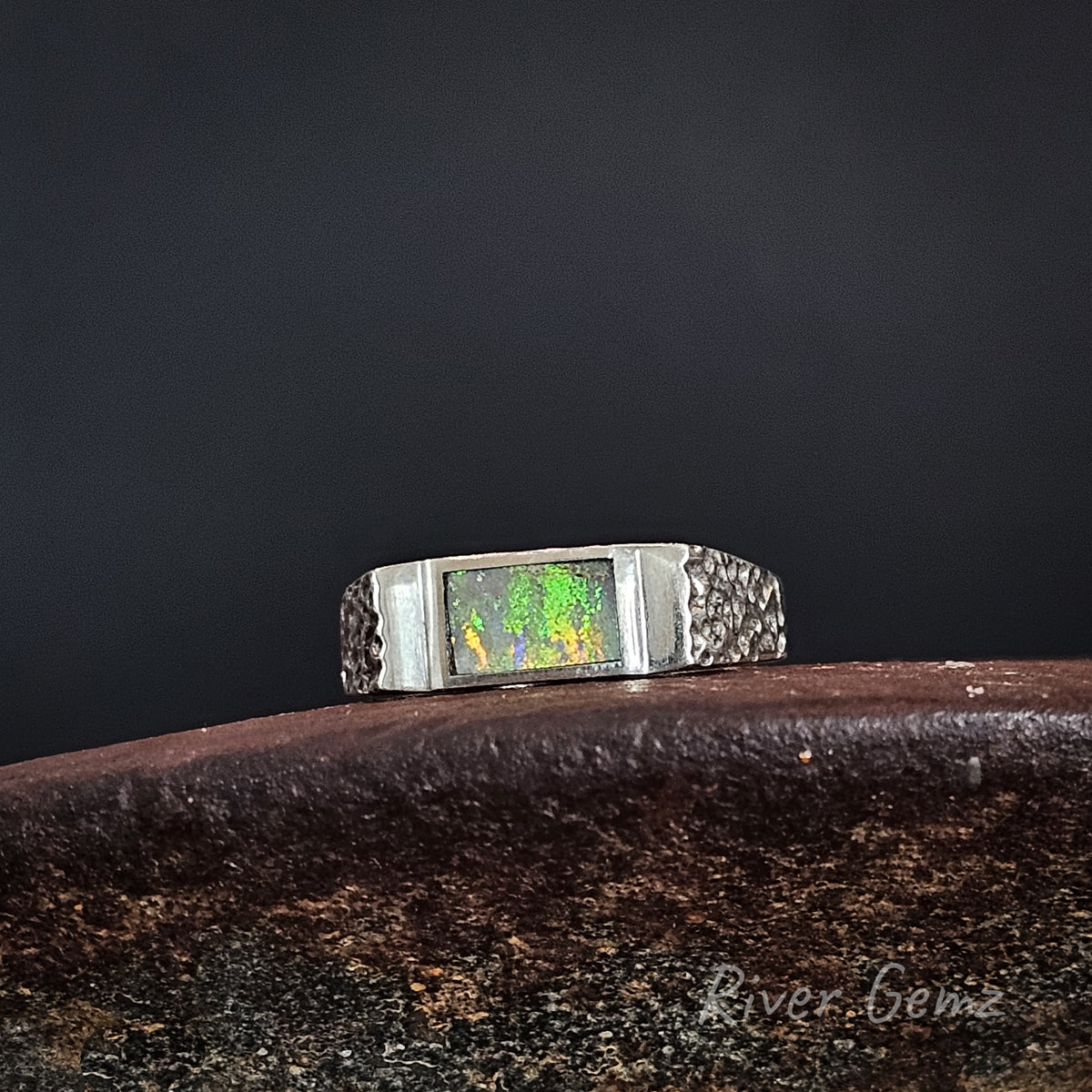 A silver-toned ring with a rectangular iridescent opal inlay, displayed on a dark textured surface against a dark background.