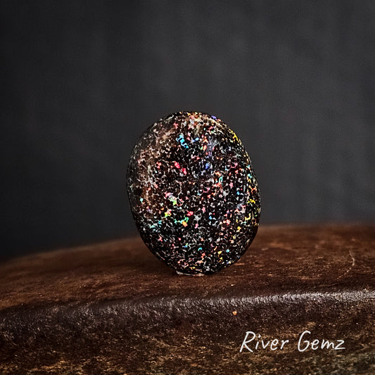 Oval shaped multi-coloured speckled matrix opal on a rusty surface branded with "River Gemz".