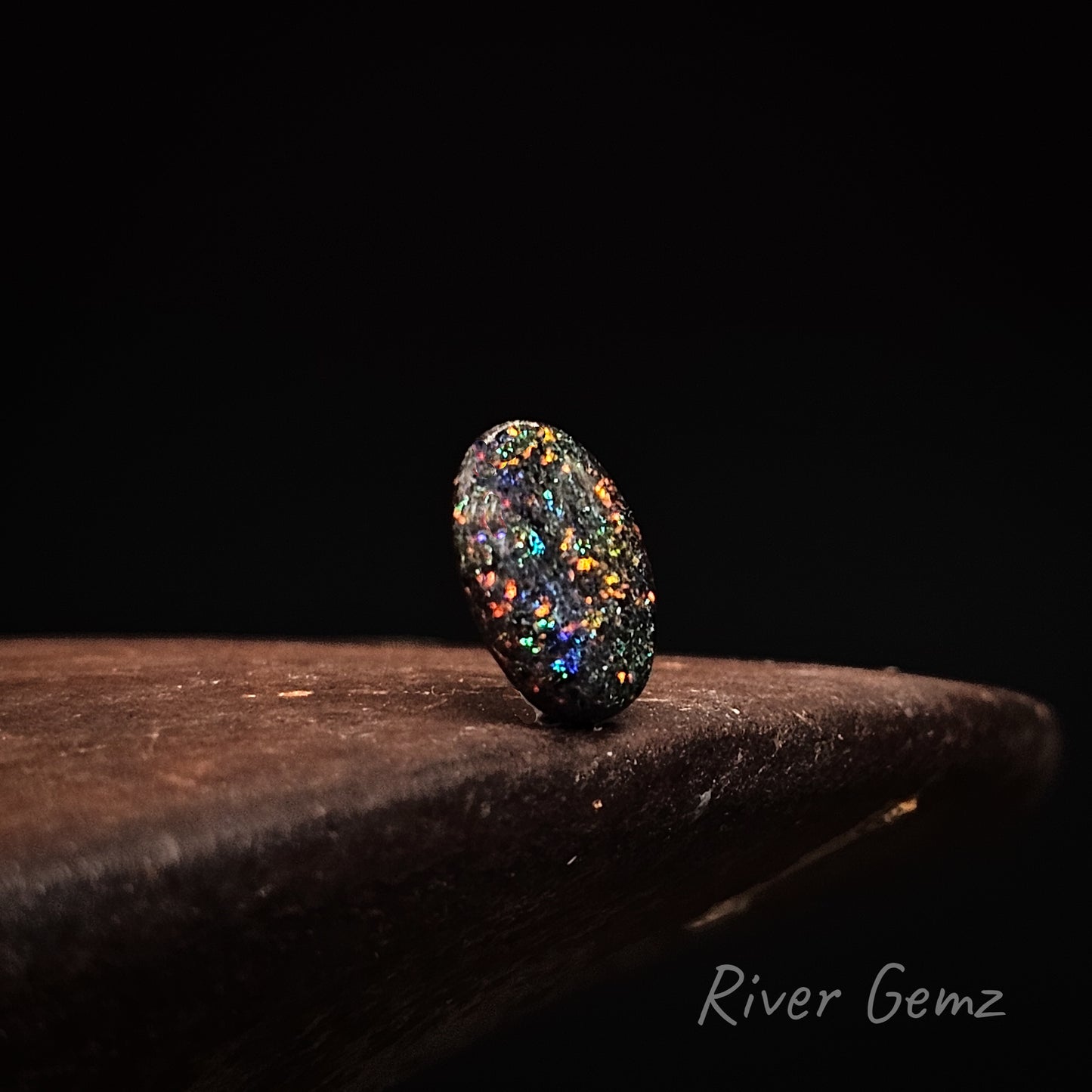 Multicolored speckled stone on a dark surface with 'River Gemz' branding.
