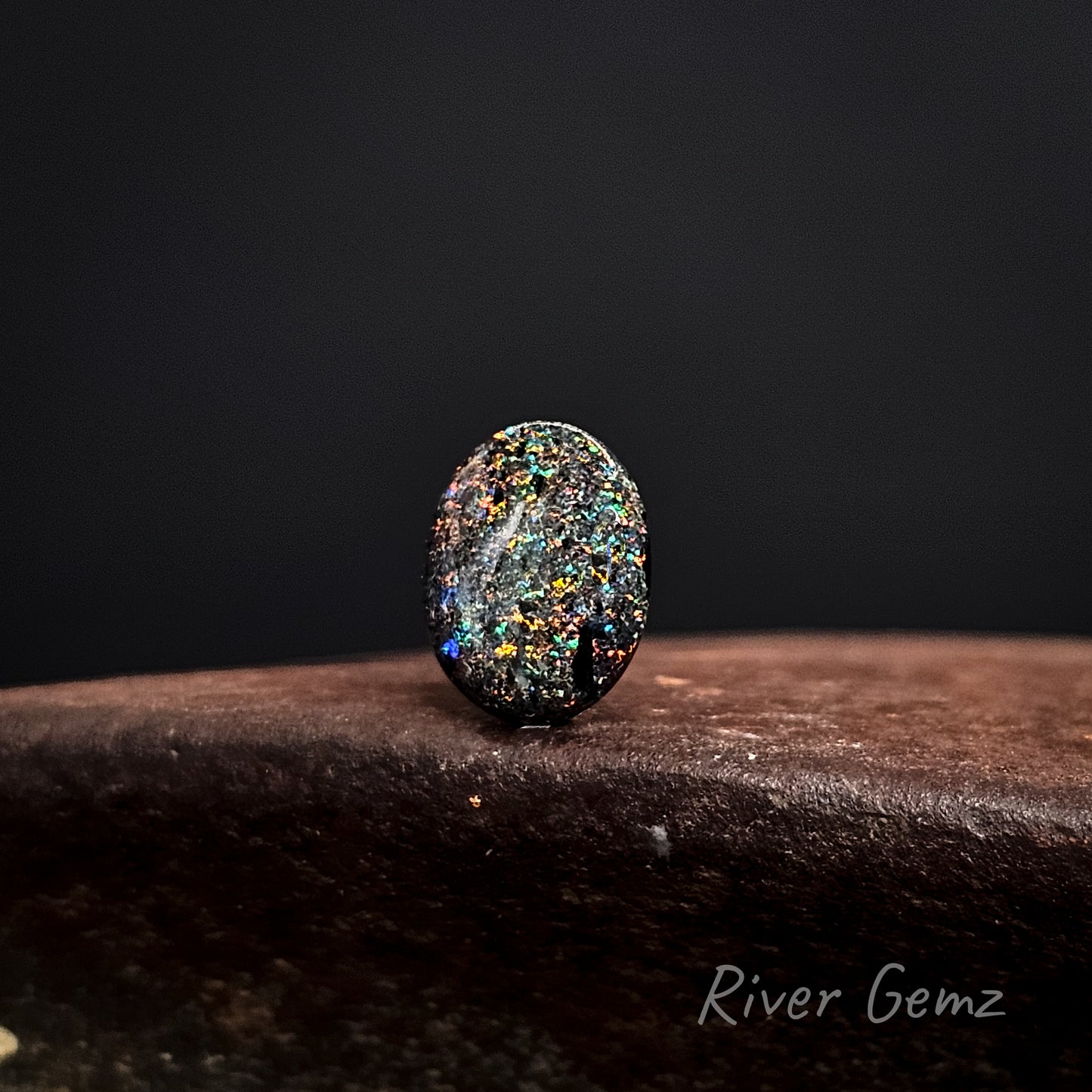 Oval shaped multicolored matrix opal on a dark background with 'River Gemz' branding.