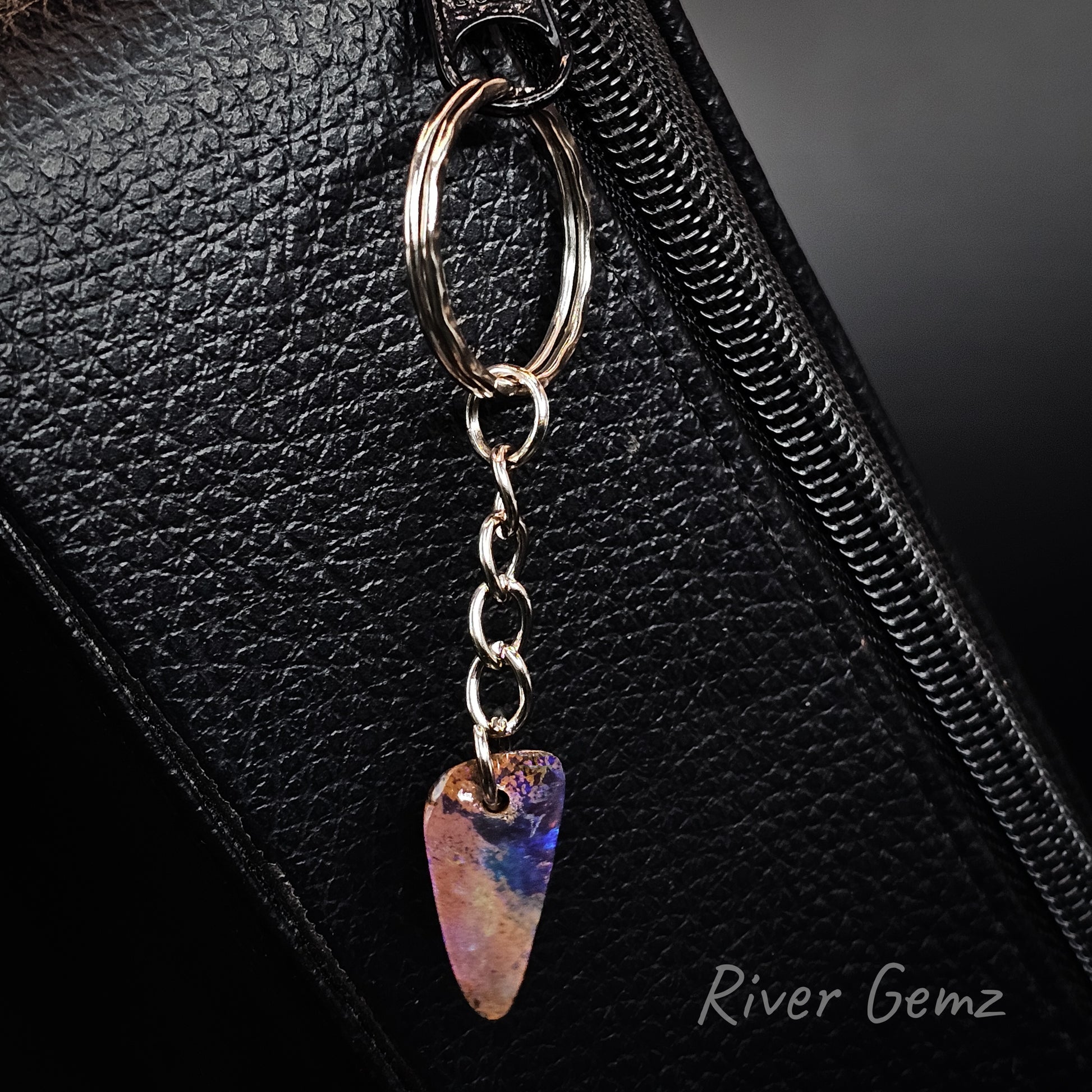 A close-up of a silver metal bag charm with a dangling iridescent boulder opal shaped like a guitar pick, resting on a black textured leather surface. The brand name 'River Gemz' is visible in the bottom right corner.