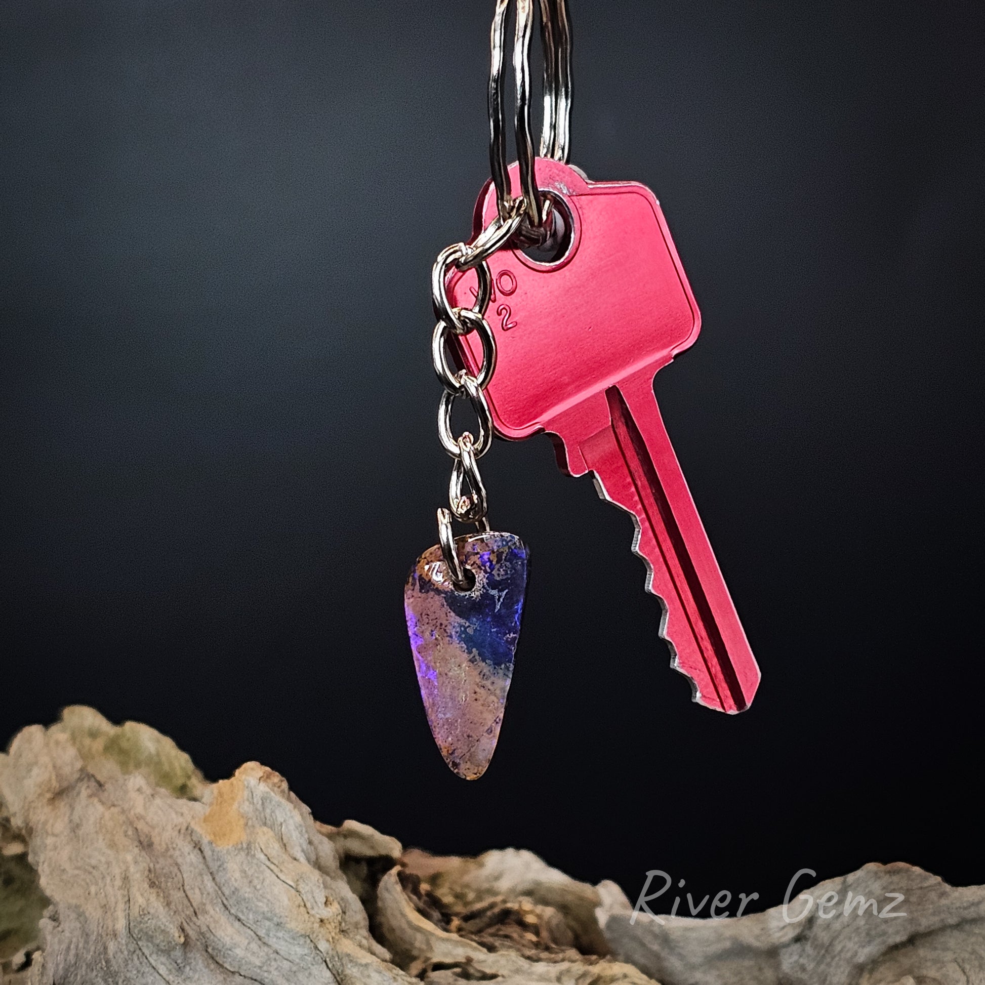 A keychain with a bright pink metal key and a dangling teardrop-shaped gemstone charm showing purple, blue, and orange inclusions, displayed against a dark background with driftwood at the bottom.
