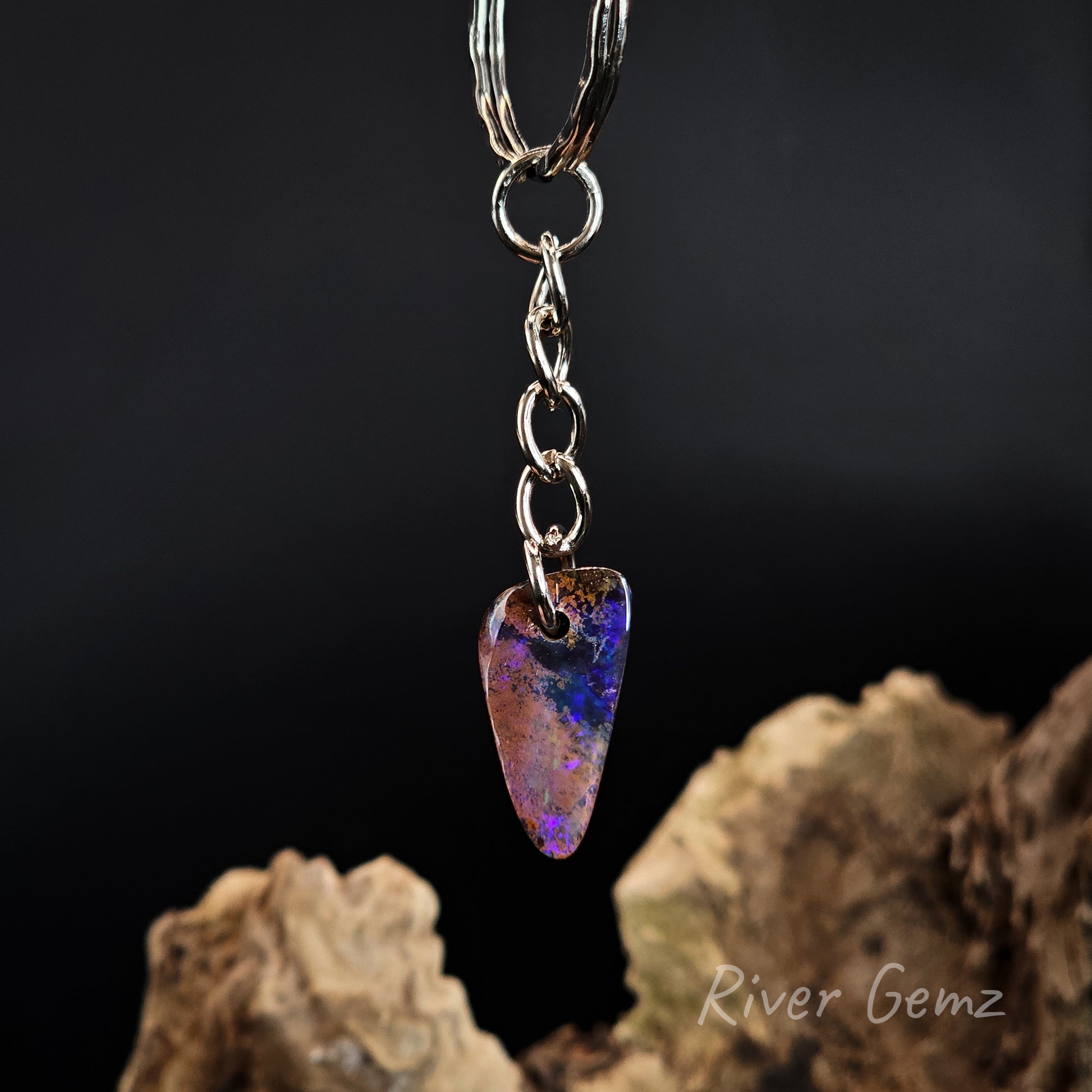 A silver-tone keychain with a teardrop-shaped iridescent opal gemstone pendant, shown hanging against a dark background with driftwood props. The gemstone displays purple, blue, and orange play-of-color.