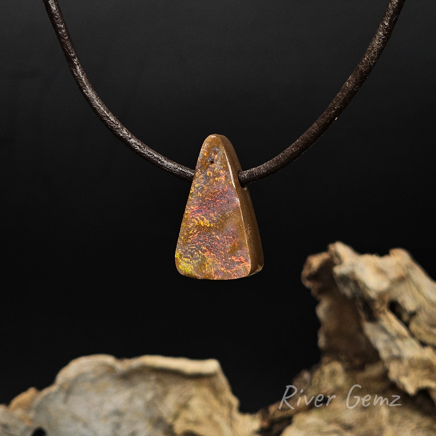 Multi-colour boulder opal on a leather cord with a black background and drift wood at the base with 'River Gemz' watermark.