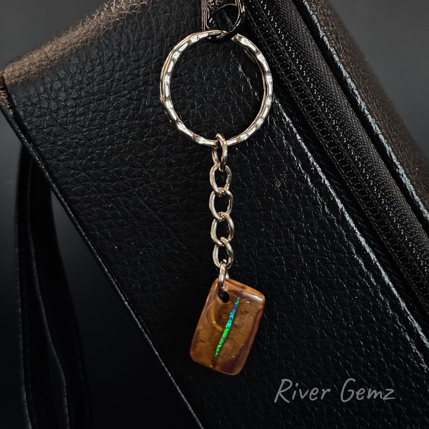 A close-up of a silver metal keychain with a textured round ring, short chain, and a rectangular brown gemstone charm featuring a vibrant green inlay. The keychain is resting on a black textured leather surface, with the brand name 'River Gemz' printed in white in the bottom right corner.