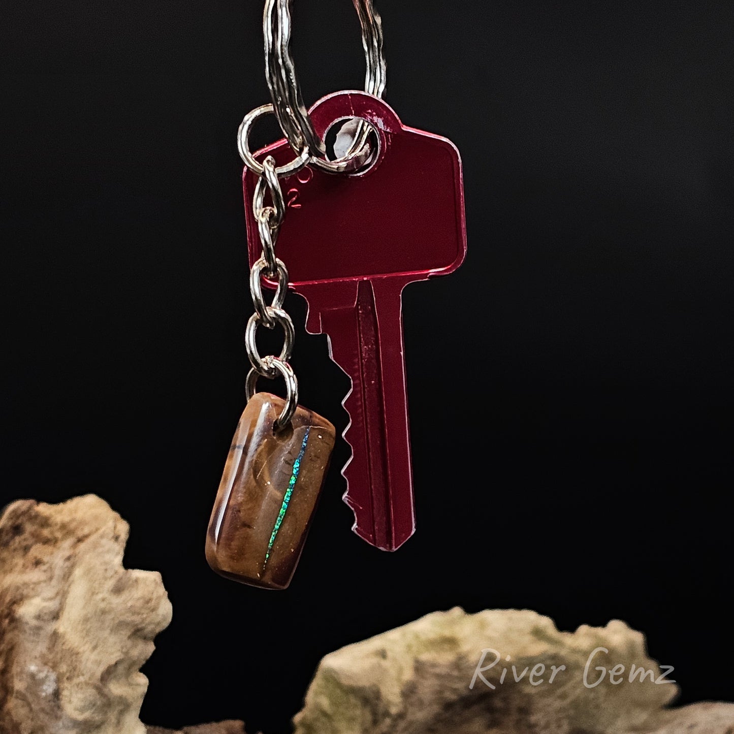 A close-up of a keychain with a small rectangular brown gemstone charm featuring a green iridescent streak, shown against a black background with rocks at the bottom. The text 'River Gemz' appears in the lower right.
