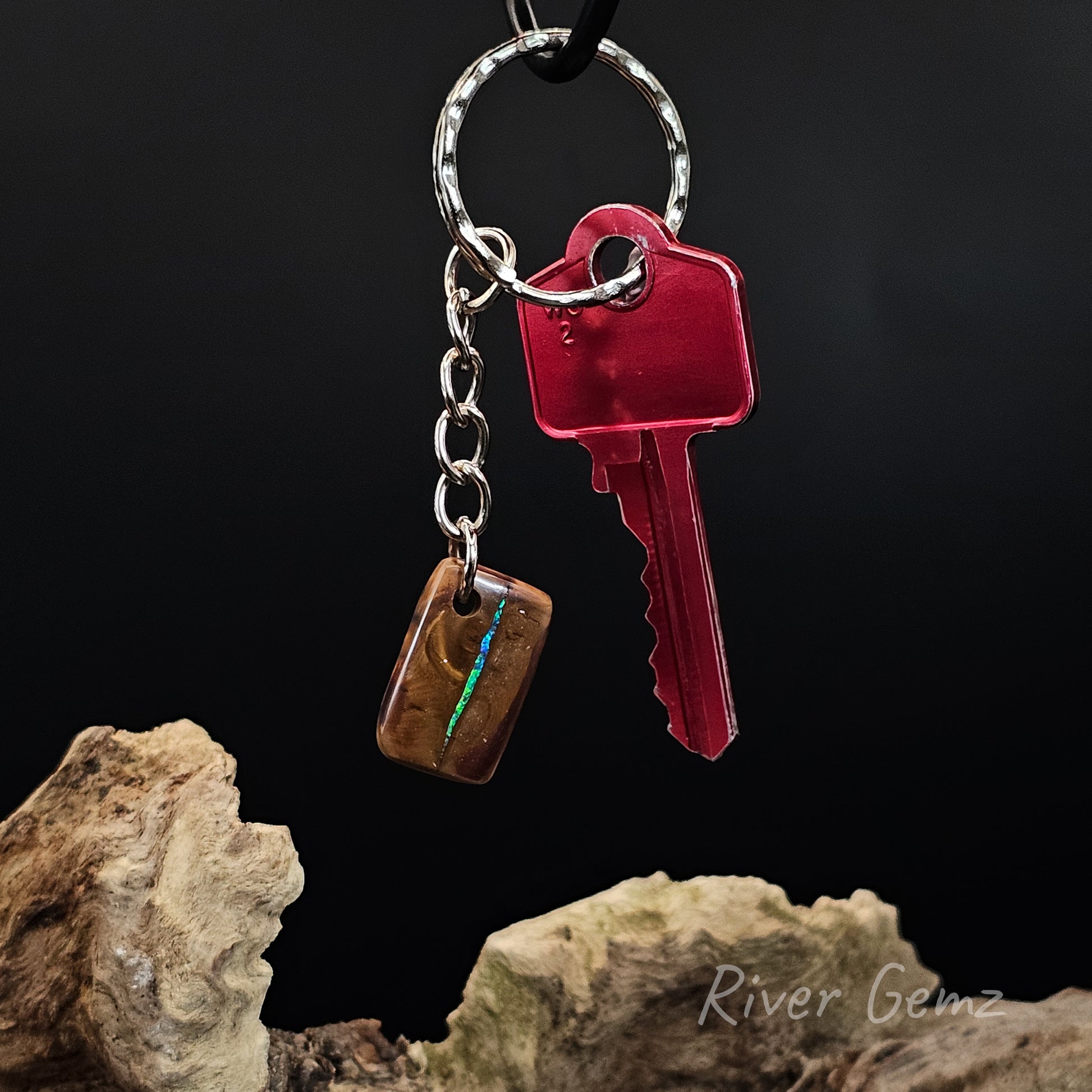 A keychain with a silver metal split ring and chain holding a red translucent key and a small brown rectangular charm with an iridescent blue-green inlay, photographed against a black background with driftwood props and the text 'River Gemz' in the lower right.