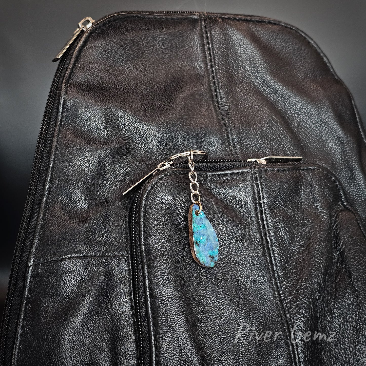 Close-up of a black leather backpack with a front zip pocket and a teardrop-shaped blue opal pendant charm on a silver zipper pull chain. The brand name 'River Gemz' is watermarked in the bottom right corner.