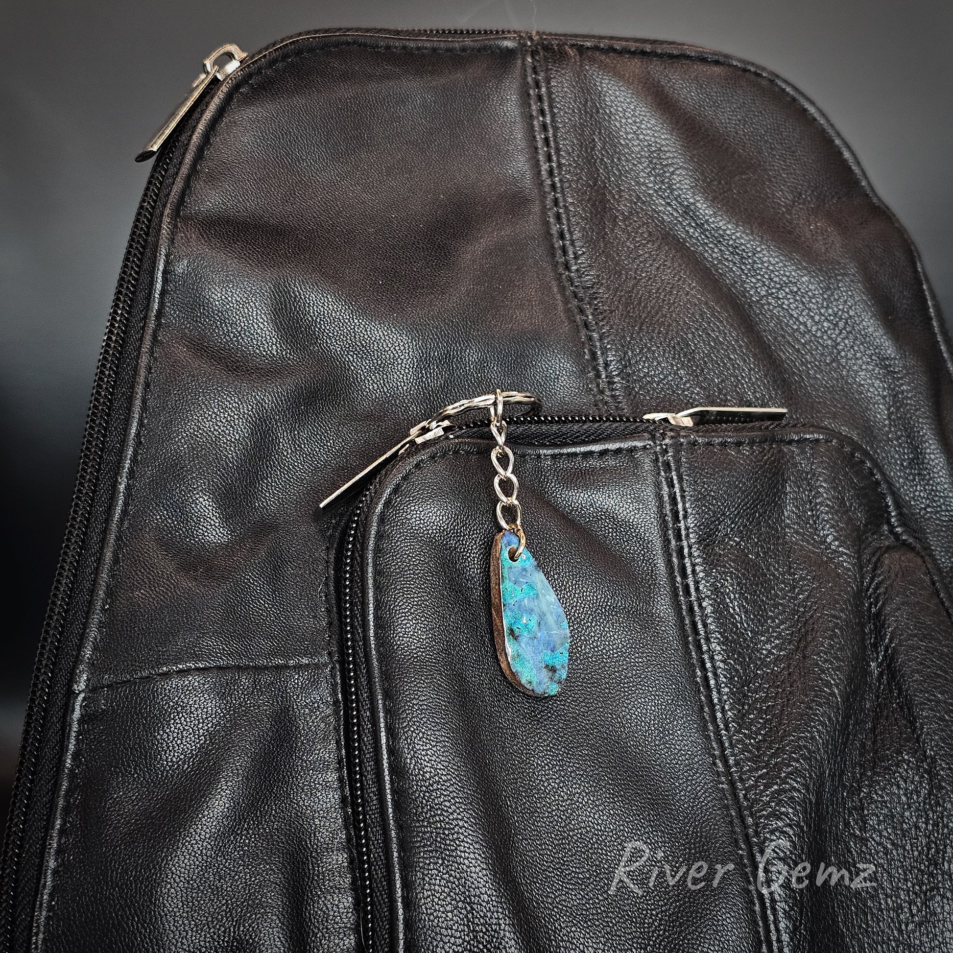 Close-up of a black leather backpack with a front zip pocket and a teardrop-shaped blue opal pendant charm on a silver zipper pull chain. The brand name 'River Gemz' is watermarked in the bottom right corner.