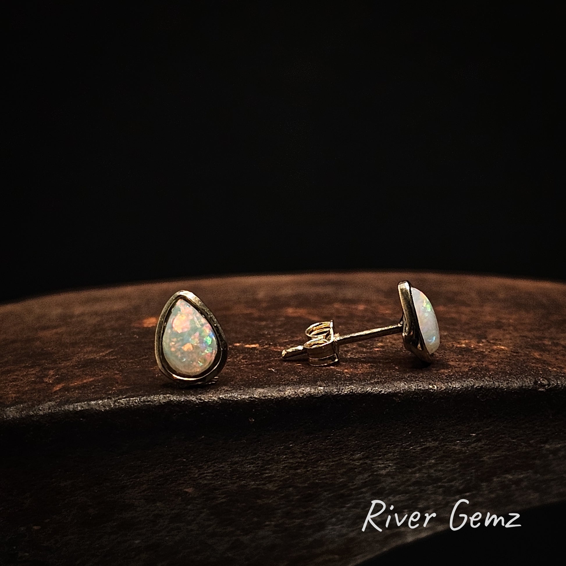 A front view and a side view of a pair of gold and opal earring studs on a dark background with 'River Gemz' branding.
