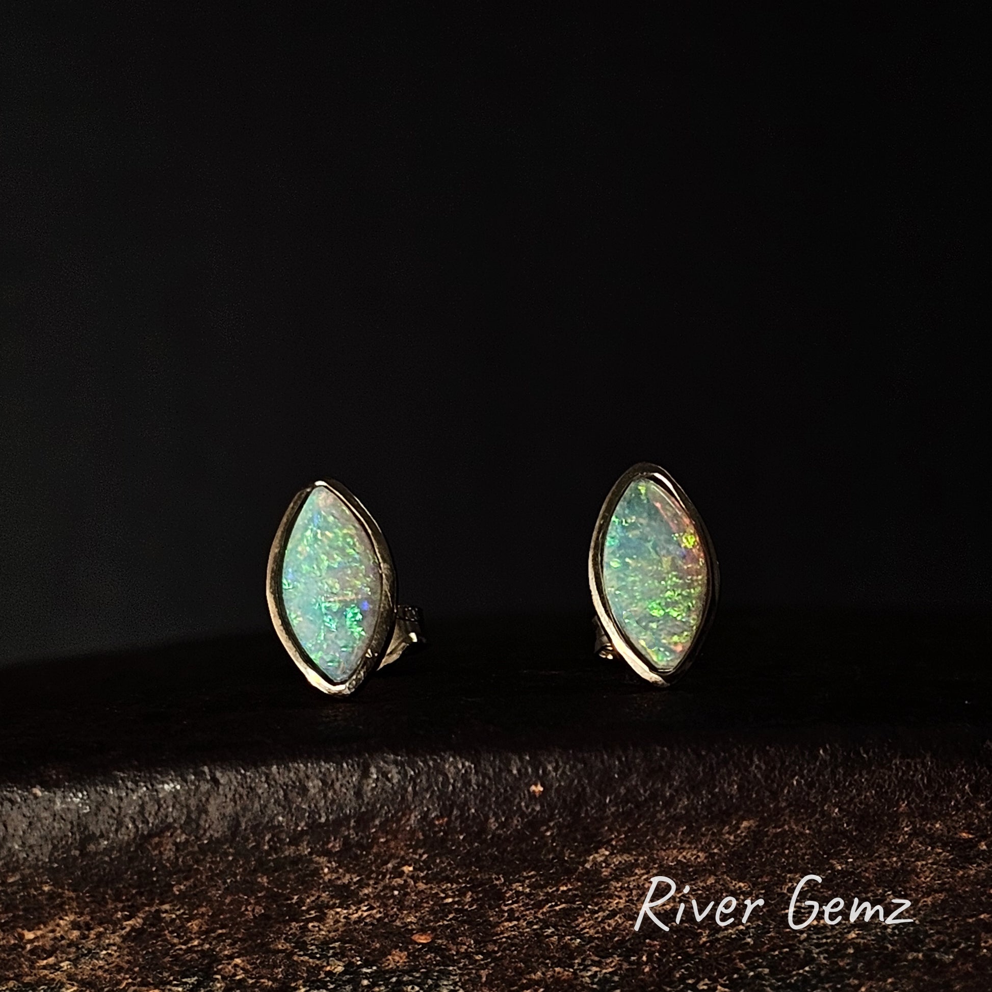 A pair of gold studs with besel set crystal opals against a dark background.